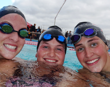 three female swimmers in a pool smiling with caps and goggles.